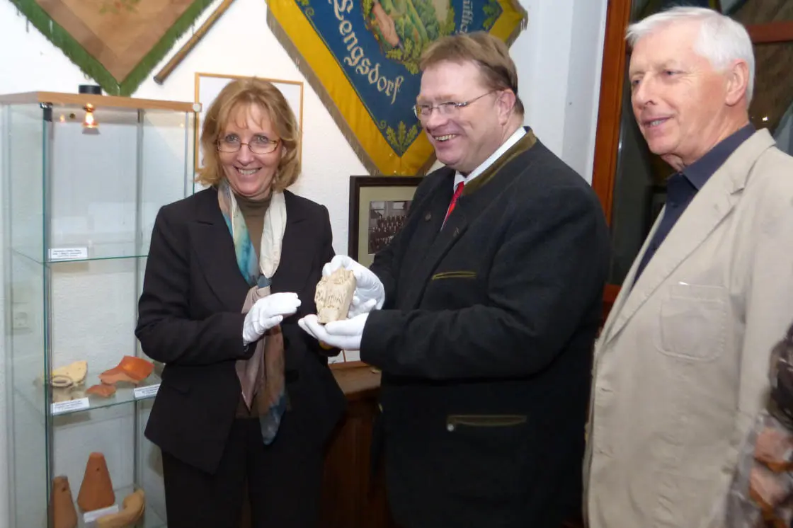 Drei Personen stehen im Heimatmuseum Lengsdorf. Eine Frau mit Brille und Schal (links) und ein Mann im Janker mit roter Krawatte (Mitte) halten gemeinsam mit weißen Schutzhandschuhen ein archäologisches Fundstück. Rechts steht ein älterer Mann im hellen Jackett. Im Hintergrund eine Vitrine mit Keramikfunden sowie die blau-goldene Vereinsfahne des HVV Lengsdorf.