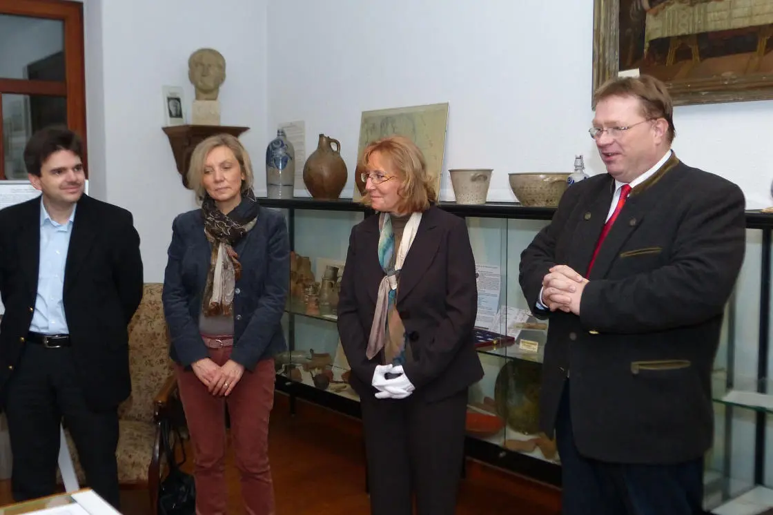 Vier Personen stehen nebeneinander im Ausstellungsraum des Heimatmuseums Lengsdorf. Rechts spricht ein Mann im Janker mit roter Krawatte zu den Gästen. Von links: ein jüngerer Mann im weißen Hemd, eine Frau mit dunklem Jackett und gemustertem Schal, eine Frau mit Brille und beigem Schal. Im Hintergrund eine Vitrine mit Keramikgefäßen und archäologischen Fundstücken, eine Büste auf einem Wandregal sowie ein gerahmtes Gemälde.