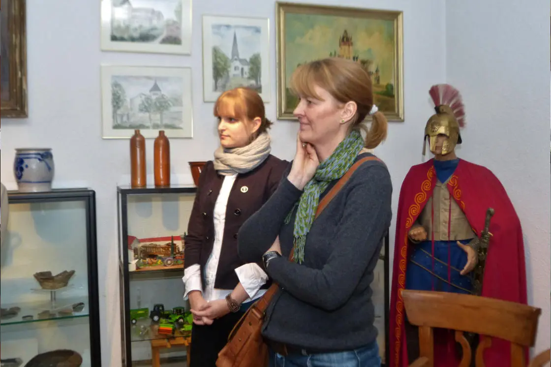 Zwei Frauen besichtigen den Ausstellungsraum des Heimatmuseums Lengsdorf. Die vordere Frau mit grünem Schal und Umhängetasche schaut nachdenklich nach rechts. Hinter ihr steht eine jüngere Frau mit hellem Schal. Rechts im Bild eine lebensgroße Figur eines römischen Legionärs mit Helm und rotem Umhang. Im Hintergrund Vitrinen mit Exponaten, Keramikgefäße sowie mehrere gerahmte Gemälde mit Lengsdorfer Ortsansichten.