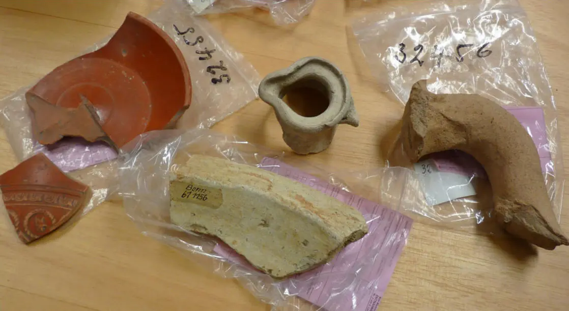 Fünf archäologische Keramikfragmente liegen auf einer Holzfläche, teils in beschrifteten Klarsichtbeuteln. Links: zwei rote Terra-Sigillata-Scherben mit Reliefdekor und Inventarnummer 33453. Mitte: ein kleiner grauer Miniaturkrug. Rechts: ein großes beiges Keramikfragment — ein Amphorenhenkel — mit Inventarnummer 32456. Im Vordergrund: eine flache helle Scherbe mit Aufkleber „Bonn 61 M56".