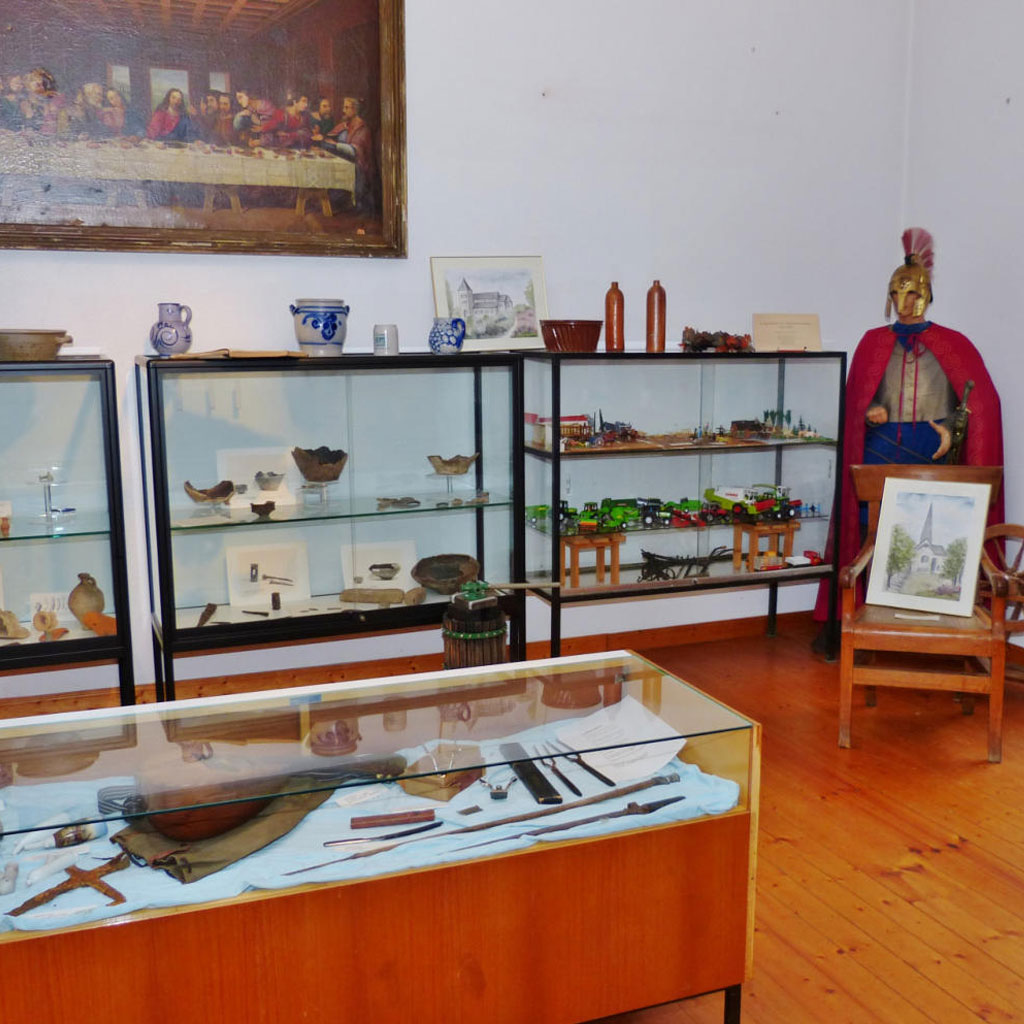 Ausstellungsraum des Heimatmuseums Lengsdorf mit mehreren Vitrinen und freistehenden Exponaten. Links zwei hohe Glasvitrinen mit archäologischen Funden und Keramikgefäßen. Im Vordergrund eine flache Tischvitrine mit Metallgegenständen, beschriftet mit „NEU". Rechts eine lebensgroße Figur eines römischen Legionärs mit Helm und rotem Umhang. Daneben ein Holzstuhl mit Aquarell, ein Spinnrad und eine Kommode. An der Wand oben links ein großformatiges Gemälde des Letzten Abendmahls.