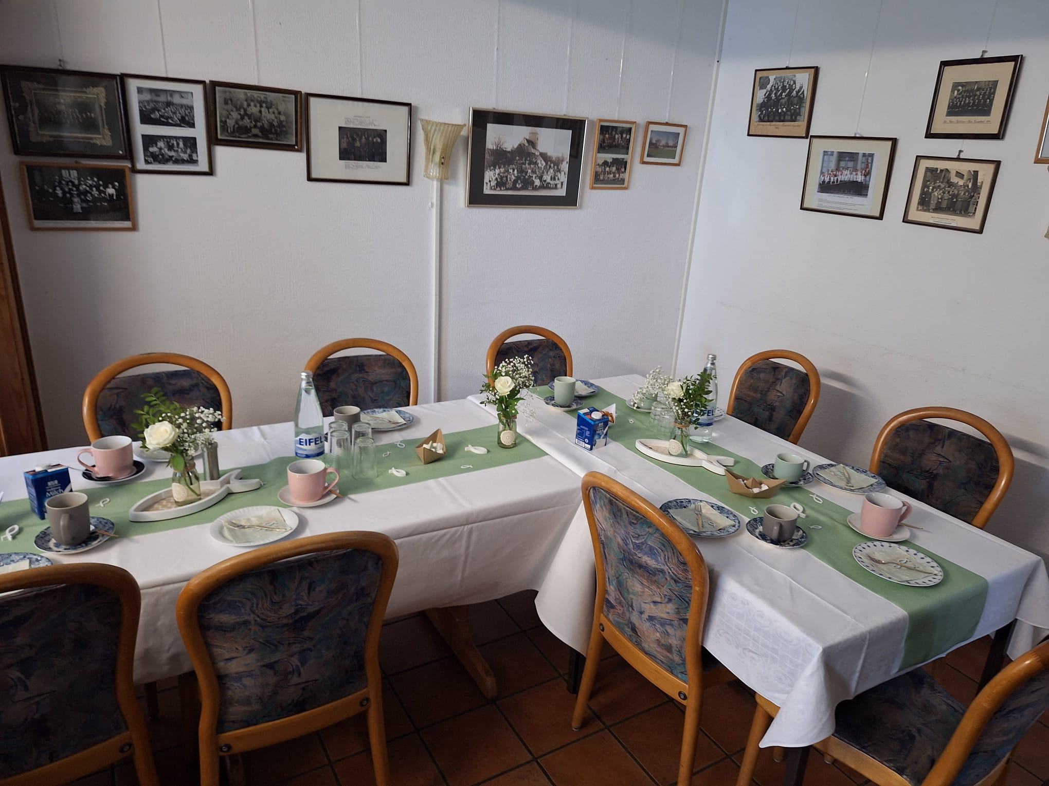 Tische3 Zwei gedeckte Tafeln im Seminarraum mit weißen Tischdecken, grünen Tischläufern, Blumenschmuck und Kaffeegedeck, an den Wänden gerahmte historische Gruppenfotos