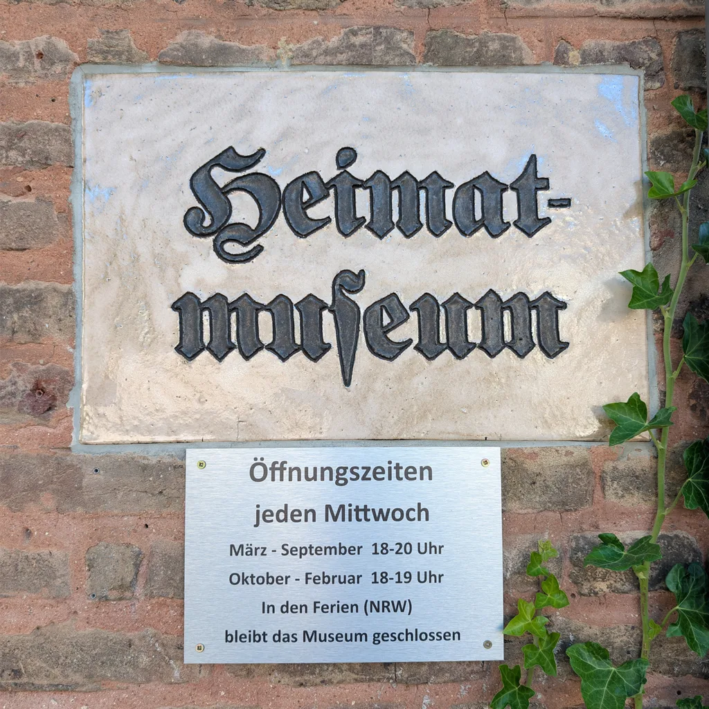Schild Heimatmuseum in Frakturschrift an einer Ziegelwand, darunter eine Tafel mit den Öffnungszeiten, rechts Efeu